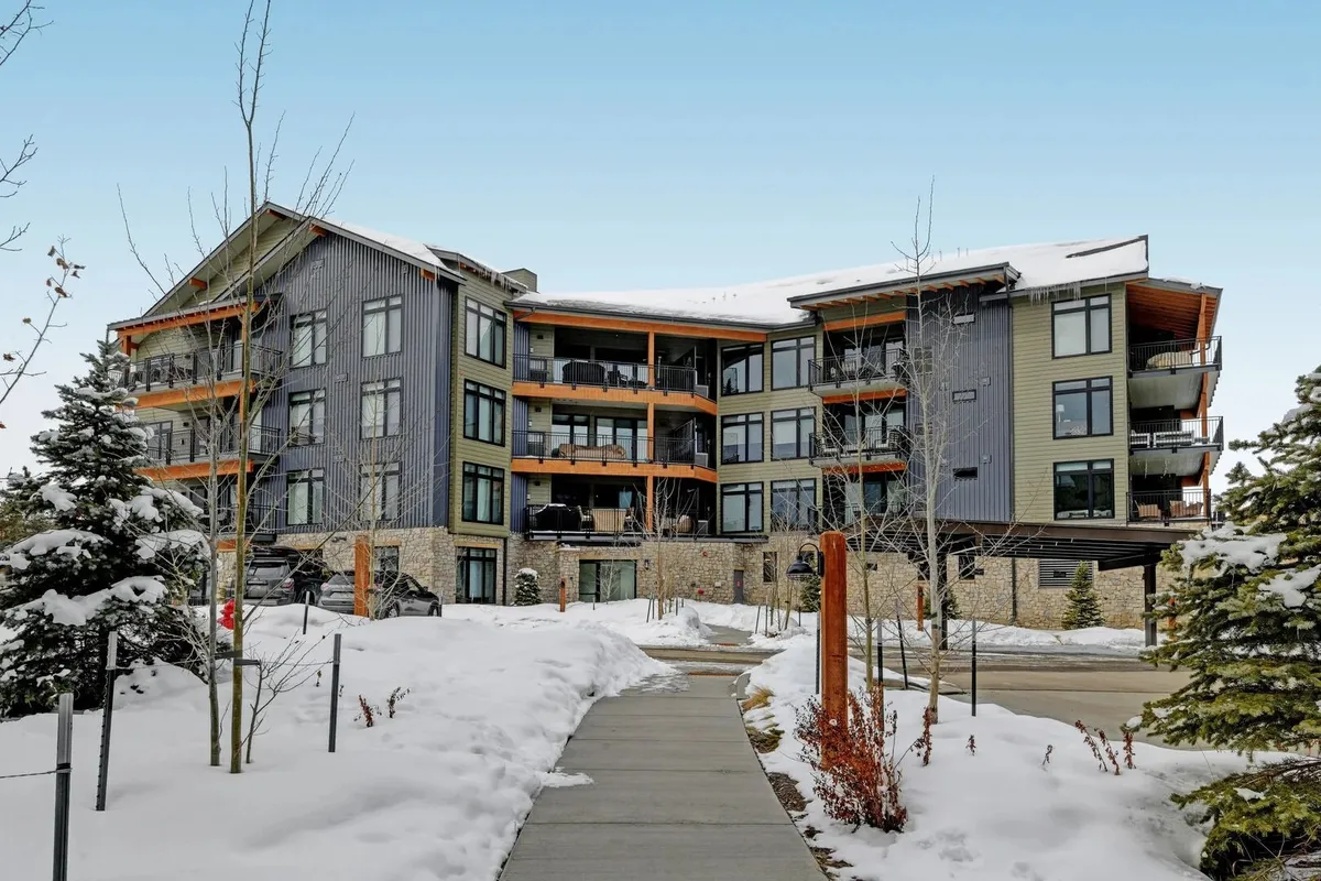 The entrance to the condo building is shown, framed by modern architecture, snow-covered pathways, balconies, and trees. The setting is cozy and welcoming, suggesting easy access to both relaxing indoors and all the downtown excitement just beyond.