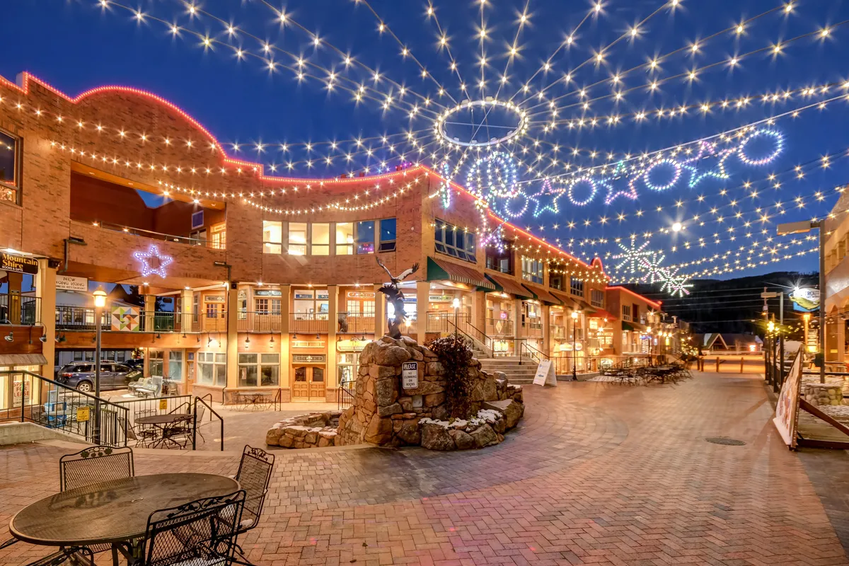 A cheerful downtown streetscape illuminated with twinkling string lights, a central stone fountain, inviting outdoor seating, and decorative storefronts. The setting suggests lively social gatherings, shopping, al fresco dining, and festive celebrations in Winter Park’s downtown core.