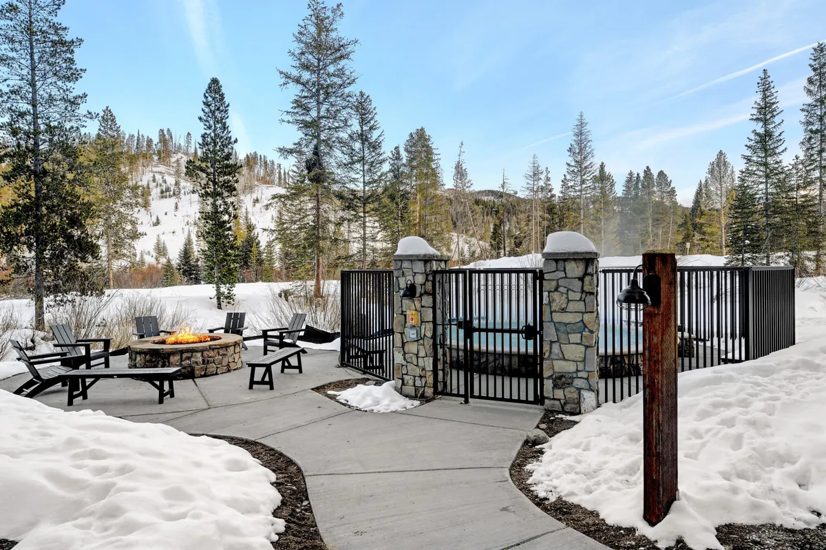 An inviting outdoor scene featuring a glowing fire pit surrounded by chairs, a bubbling hot tub, snow-covered ground, and a forest backdrop. The atmosphere is cozy yet invigorating, suggesting relaxation after an active day of exploration.