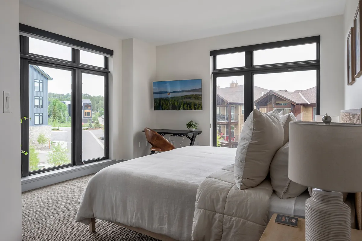 A serene bedroom featuring a plush bed with decorative pillows, large windows that let in natural light, a mounted TV, and a stylish lamp. The soft hues and gentle lighting evoke tranquility and comfort, making it an ideal space for relaxation and unwinding after a day of adventure.