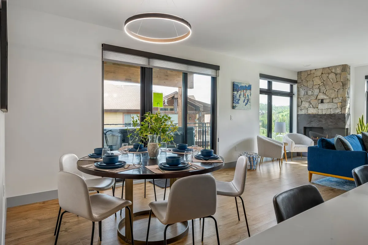 A dining area seamlessly connected to the living space, showcasing a custom wood dining table set for a meal, a stone fireplace, blue sofa, and expansive windows offering a scenic backdrop. The layout is open and ideal for gatherings.