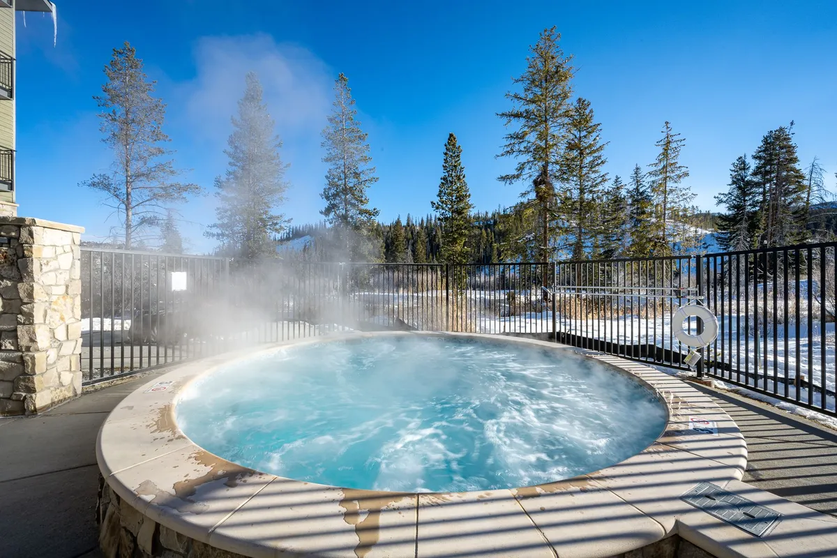 An exterior shot featuring a luxurious hot tub nestled among tall trees, with a breathtaking view of the surrounding mountains. The hot tub is the focal point, inviting guests to relax while immersed in nature. The tranquil mood is enhanced by hints of a starry night sky and the warm glow from the tub area.