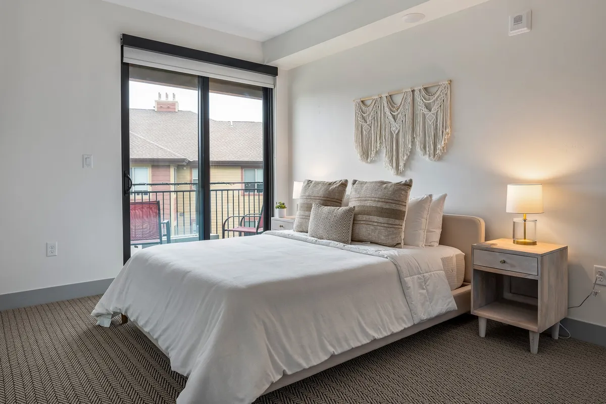 A cozy bedroom with a comfortable bed adorned with decorative pillows, a macrame wall hanging, a bedside lamp casting a gentle glow, and a view out to a private balcony. The intimate setting is perfect for relaxed mornings or quiet evenings.