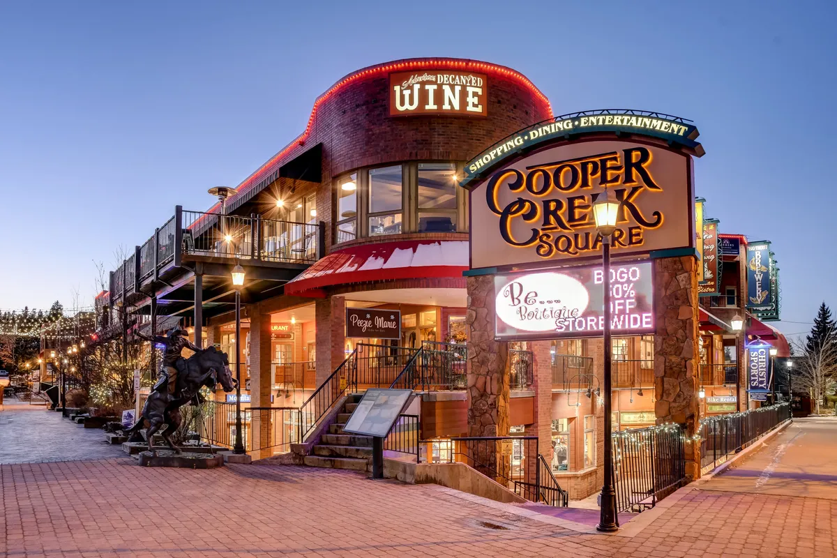 The inviting exterior of Cooper Creek Square, adorned with decorative lighting and outdoor seating. Signs for wine and dining are visible, with people mingling in a festive, welcoming atmosphere. This scene represents the vibrant, walkable downtown environment.