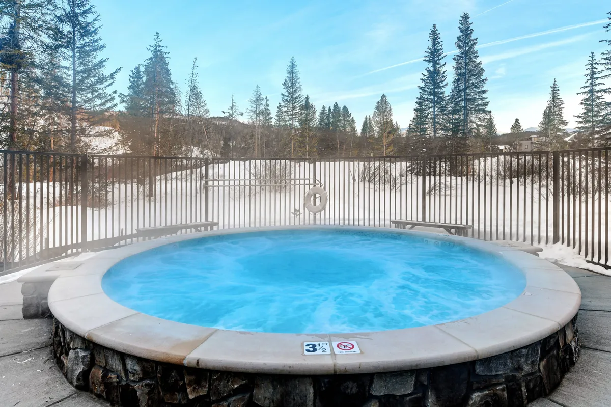 An inviting outdoor hot tub set against a snowy landscape, surrounded by trees. The warm steam rises as the sun sets behind the mountains, evoking both winter relaxation and scenic beauty.