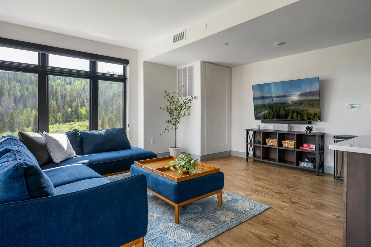 The image showcases a spacious and inviting living room with a plush blue sectional sofa and a stylish coffee table adorned with greenery, bathed in natural light from expansive windows that reveal panoramic mountain views. The space exudes comfort and warmth, perfect for gathering with friends or unwinding as the sun sets over Winter Park.