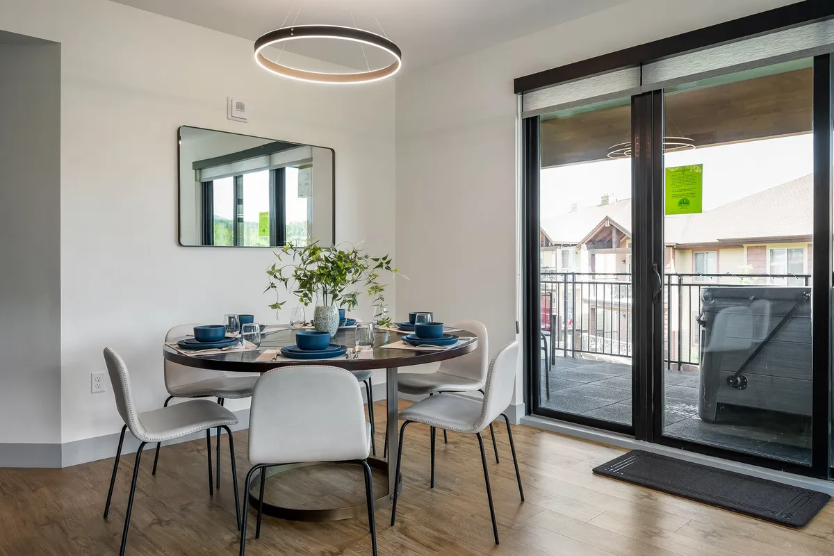 A round dining table with blue dinnerware and a plant centerpiece, positioned near large glass sliding doors that open to a private balcony. Natural light pours in, hinting at easy access to the outdoors and inviting forest views.